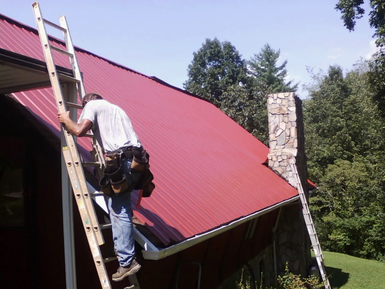 Roofer on a ladder beside a red metal roof and stone chimney — Soffit & Fascia Repair by Roofing Brothers in Lacey
